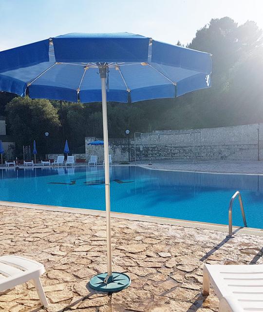 Piscina luminosa, ombrelloni blu, sdraio bianche, atmosfera serena, sole splendente, relax estivo.