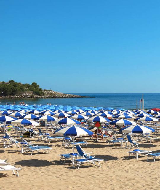 Spiaggia affollata di ombrelloni blu e bianchi, sole splendente e mare calmo all'orizzonte.