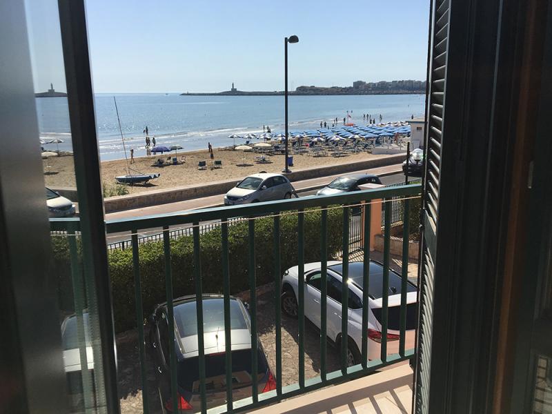 Vista mare da un balcone, spiaggia soleggiata con ombrelloni e auto parcheggiate.