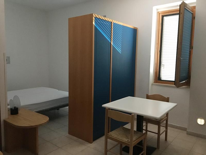 Stanza semplice e luminosa, con letto, tavolo e armadio, atmosfera tranquilla e accogliente.