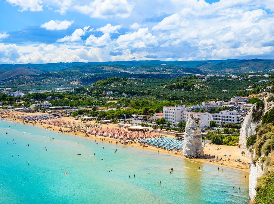 Spiaggia affollata con mare turchese, montagne verdi e cielo azzurro, atmosfera estiva e vivace.